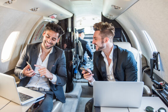 Young well dressed businessmen sitting inside private airplane and working on their mobile devices during business flight. Cockpit with pilot is seen in the background.